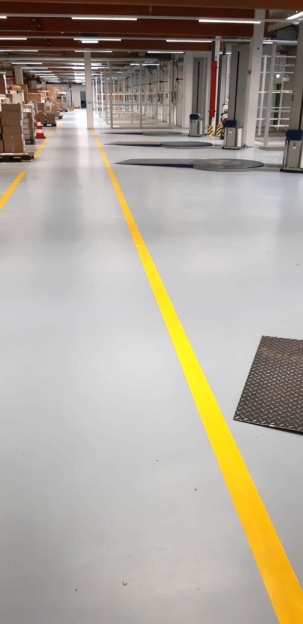 Langer Hallenkorridor mit hellgrauem Industrieboden und zwei parallelen gelben Fahrspurmarkierungen – Lagerbereich mit Regalen und Packstationen