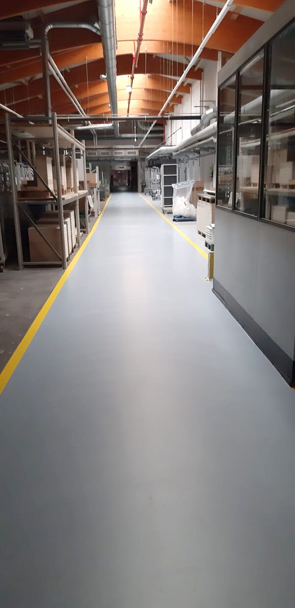 Langer Hallengang mit hellgrauem Epoxidharz-Boden und gelber Fahrbahnmarkierung – Produktionshalle mit orangefarbenem Bogendach