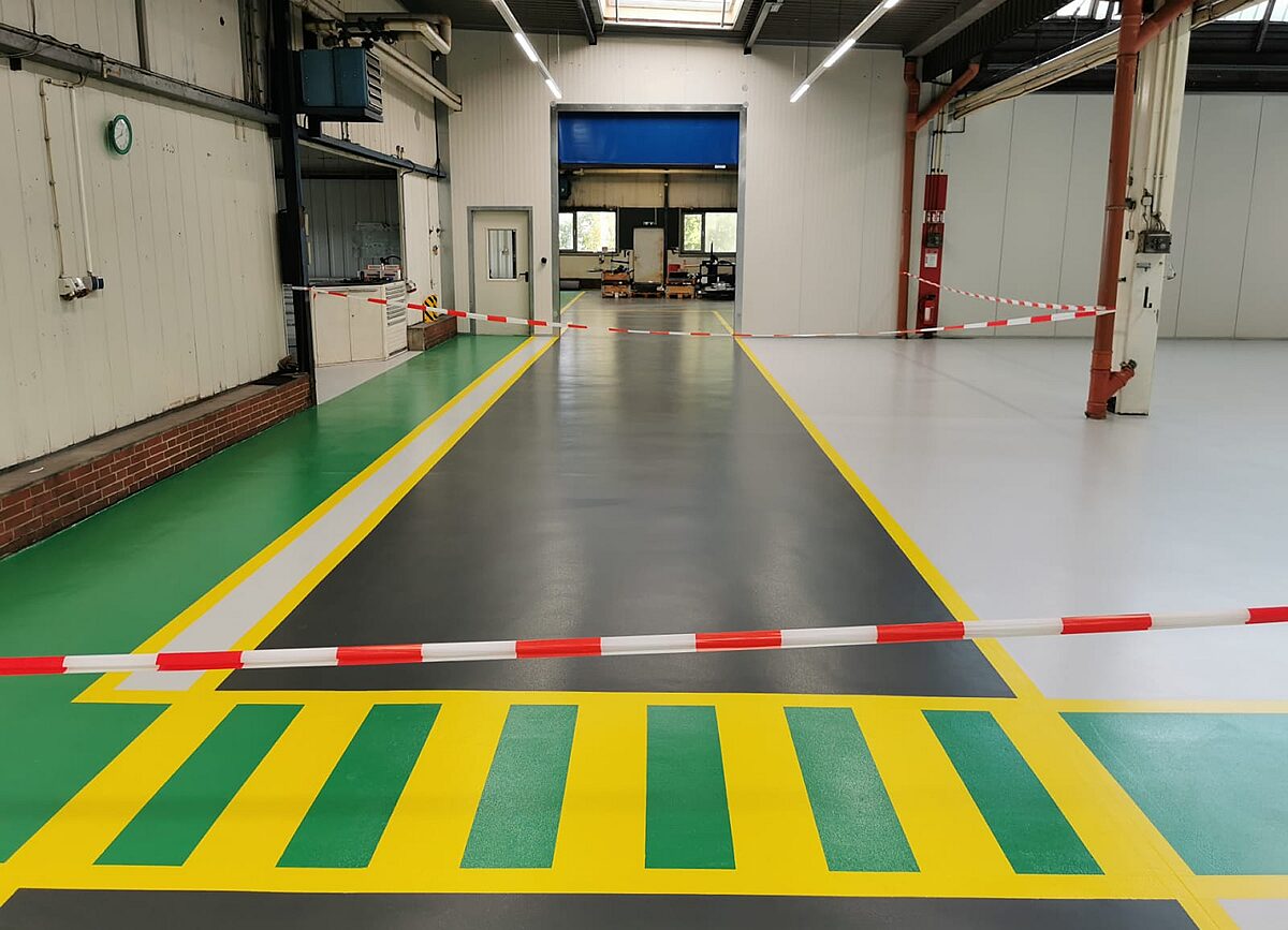 Dekorative mehrfarbige Industrieboden-Beschichtung – grüne Fußgängerfläche, gelbe Trennlinien und dunkelgrauer Fahrweg in einer Produktionshalle