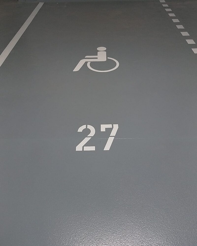 Behindertengerechter Stellplatz Nr. 27 mit weißem Rollstuhlsymbol auf dunkelgrauem Industrieboden – Tiefgarage mit gepunkteter Begrenzungslinie