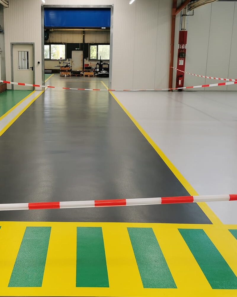 Industriehalle mit mehrfarbiger Bodenmarkierung – grüner Fußgängerüberweg mit gelbem Rahmen und dunkelgrauem Fahrweg