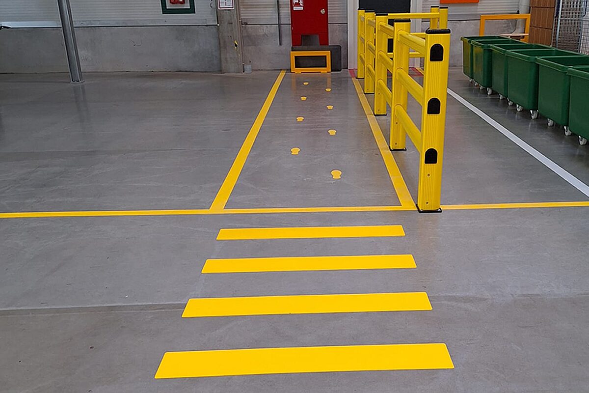 Gelbe Fahrbahnmarkierung mit Fußgängerüberweg und Laufweg neben gelbem Rammschutz-Geländer in einer Industriehalle