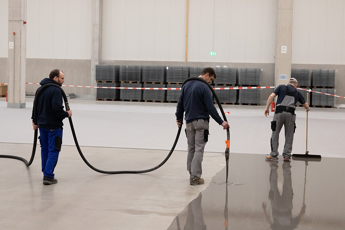 Drei Handwerker tragen mineralische Industrieboden-Beschichtung in einer neuen Lagerhalle auf – glänzende frische Bodenfläche mit Spiegelung der Hallenkonstruktion