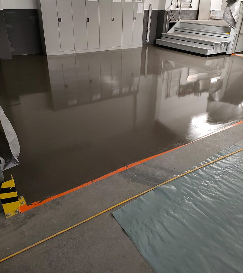 Langer Industriehallengang mit frisch aufgebrachter grauer mineralischer Bodenbeschichtung – gleichmäßige matte Oberfläche mit grünen Stahlstützen