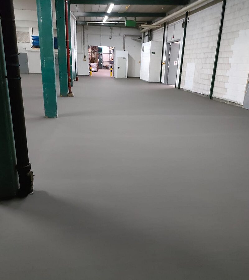 Frisch verlegte mineralische Beschichtung in einer Halle – hellgrauer glänzender Boden mit sichtbarer Verlegefront und altem Untergrund im Vordergrund