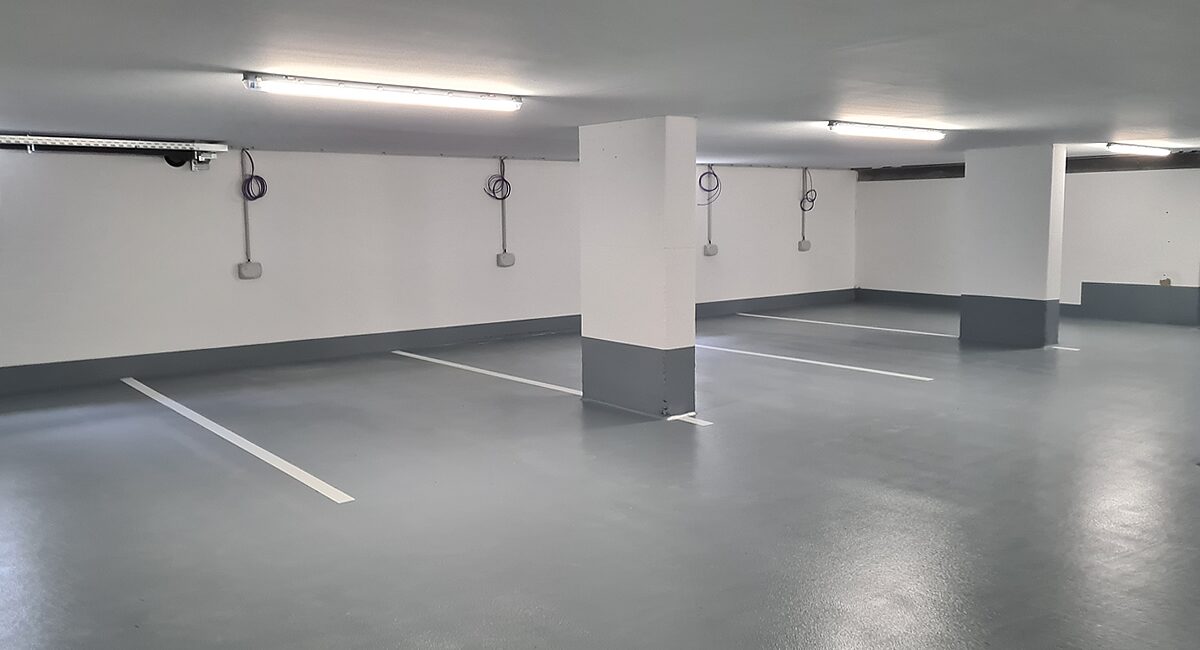 Tiefgarage mit hellgrauem OS-8-Boden – weiße Stellplatzmarkierungen, Ladesäulen an der Wand und grauer Sockelstreifen als Wandabschluss