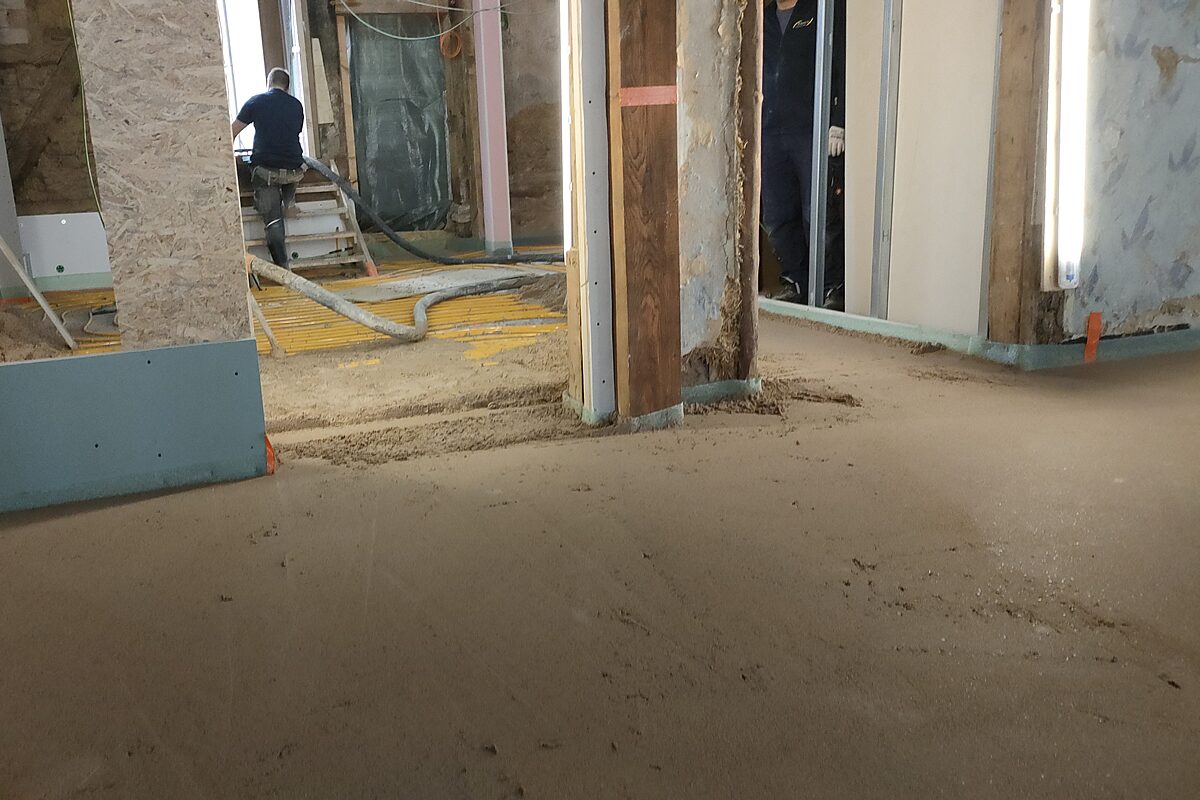 Mitarbeiter beim Estricheinbringen im Bestand Höxter – pumpt Estrichmasse mit Schlauch ein, Fußbodenheizungsrohre auf Trennlage, OSB-Trennwände und historische Holzstützen im Bild