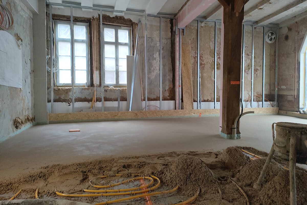 Großer Bestandsraum im Altbau Höxter vor der Estrichverlegung – freigelegte historische Wände mit Putzresten, Fachwerkstütze in der Raummitte und Fenster mit Bauschutz an den Rahmen