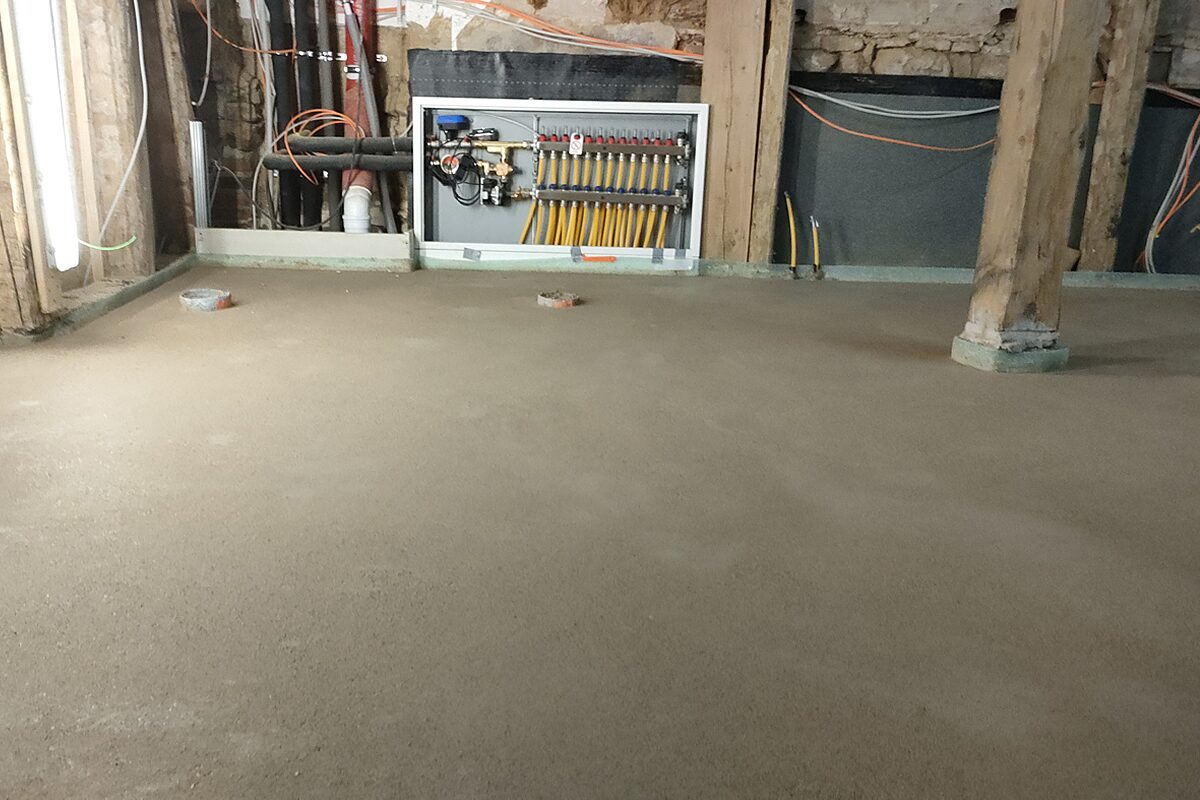 Detail im Altbau Höxter – fertig eingebrachter Estrich rund um eine historische Holzstütze, an der Wand der Heizkreisverteiler der Fußbodenheizung mit gelben und roten Stellantrieben
