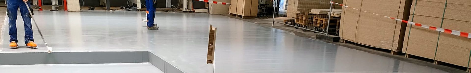 Industriebodenbeschichtung im laufenden Betrieb – zwei Mitarbeiter beim Auftragen von Epoxidharz-Beschichtung mit Rolle in einer Produktionshalle