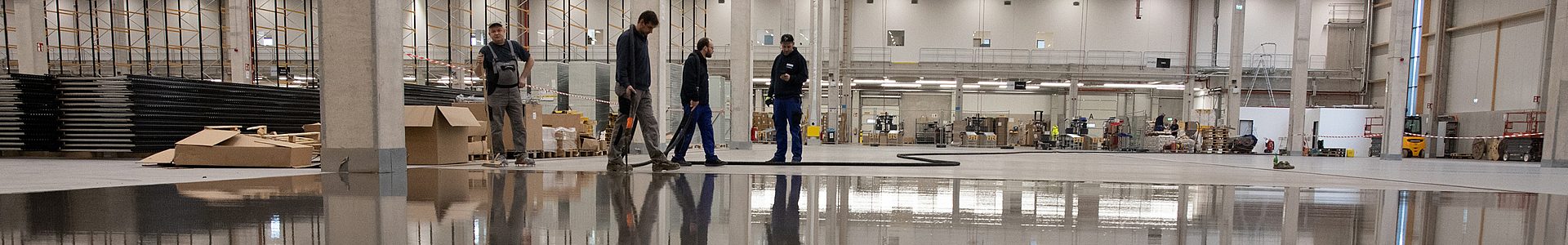 Handwerker tragen eine mineralische Beschichtung auf einem hochglänzenden Industrieboden in einer Logistikhalle auf