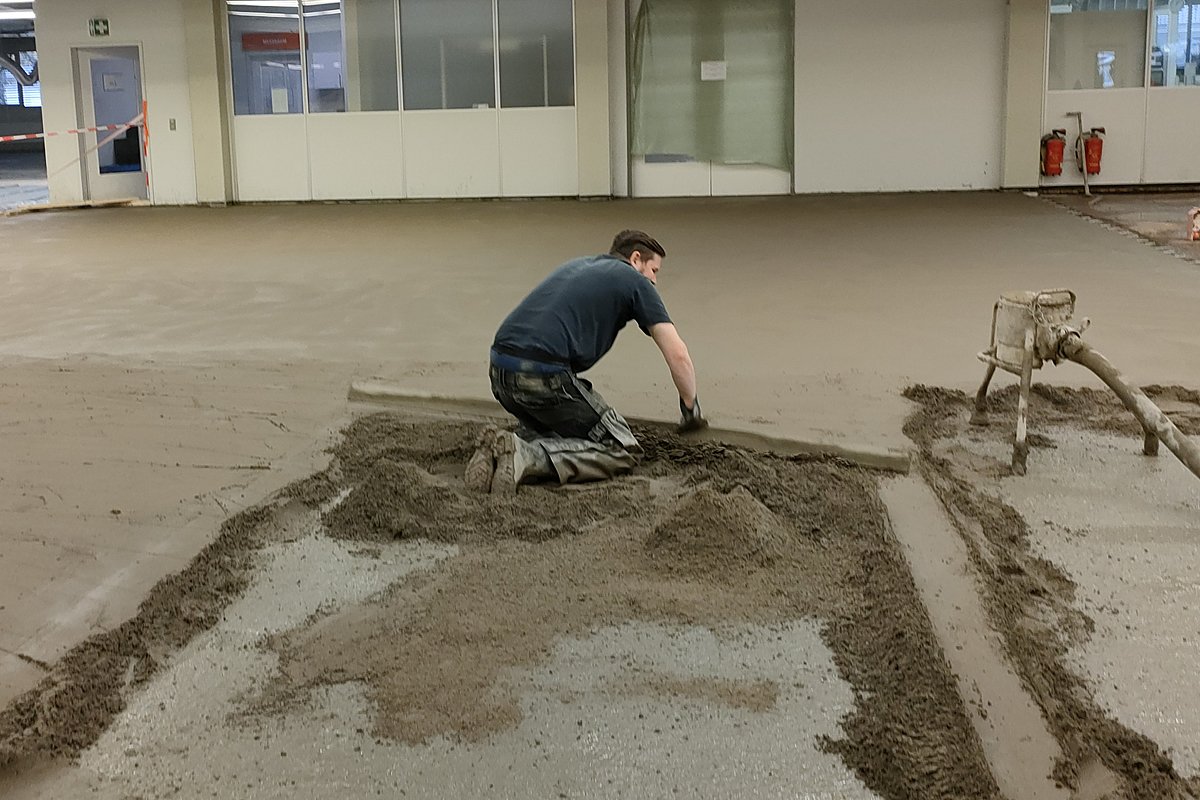 Schnellestrich Einbau – Mitarbeiter verarbeitet Zementestrich mit Kelle in großer Gewerbehalle