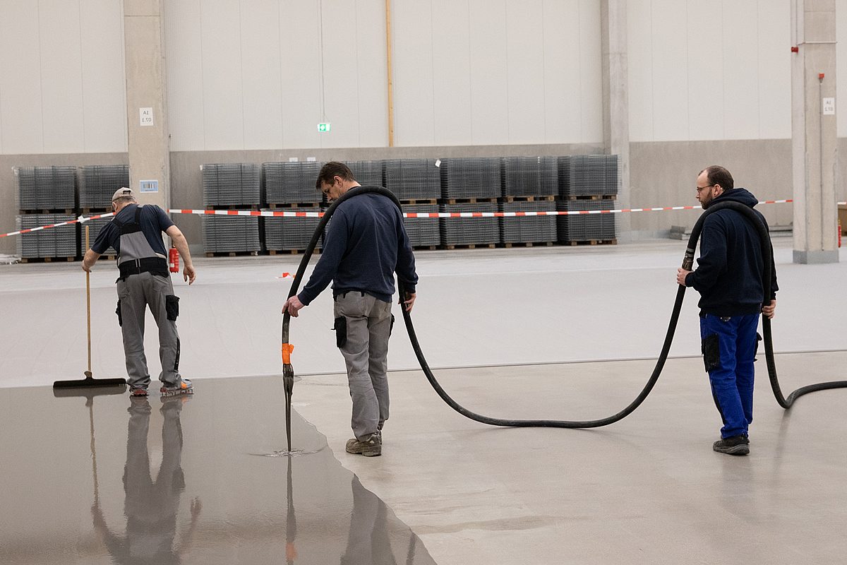 Drei Handwerker tragen Industrieboden-Beschichtung in einer neuen Lagerhalle auf – ein Mitarbeiter verteilt mit dem Rakel, zwei weitere führen den Pumpschlauch