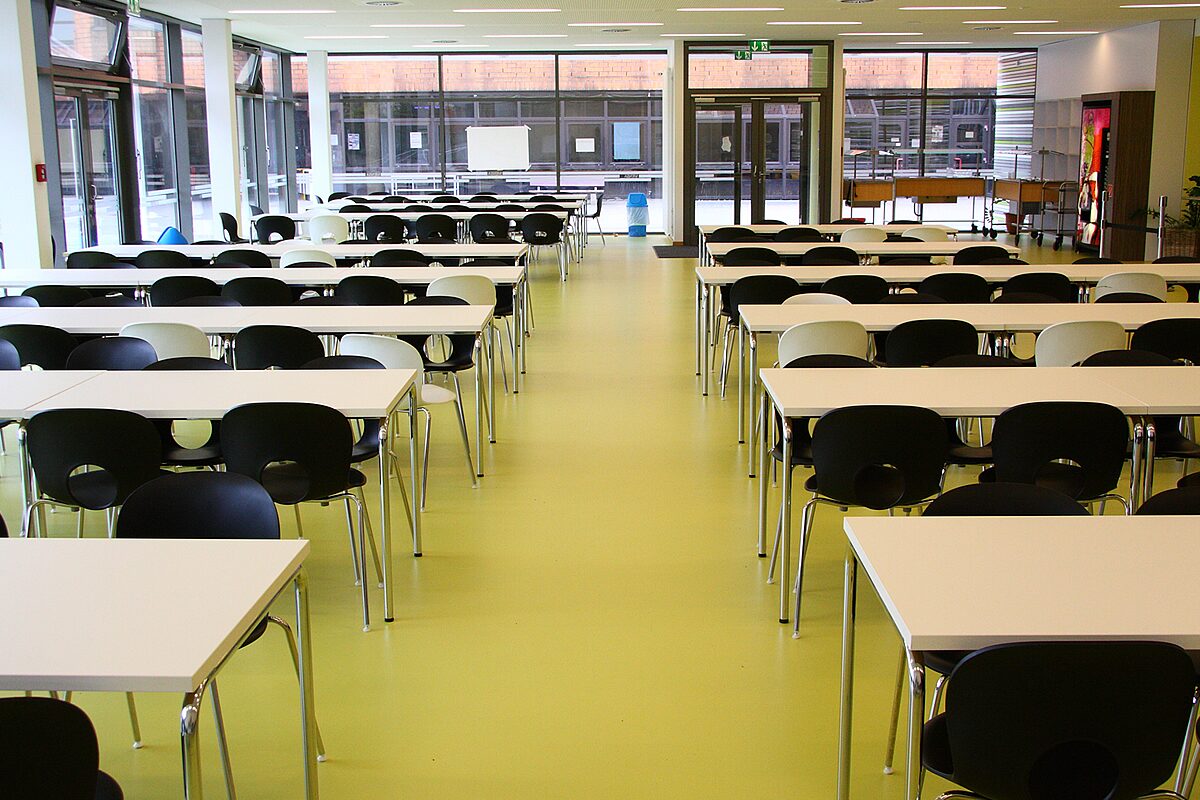 Schulkantine mit gelbem Designboden – lebhaft gefärbter nahtloser Fußboden unter weißen Tischen und schwarzen Stühlen in hellem Speisesaal